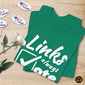 May include: A green t-shirt with the text "Links always vote" in white. The shirt is folded and lying on a wooden surface with a white rose and several "I Voted" stickers.