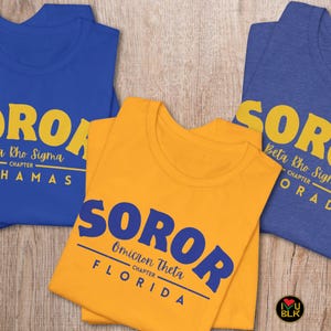 May include: Three t-shirts in blue and yellow, each with a different design. The blue shirts have "SOROR" in yellow, with text below indicating "Zeta Rho Sigma CHAPTER BAHAMAS" and "Beta Rho Sigma CHAPTER ORADO". The yellow shirt says "SOROR Omicron Theta CHAPTER FLORIDA".