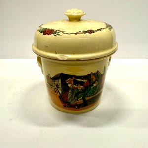 May include: A vintage, cream-colored ceramic container with a lid and two handles. The lid is adorned with a floral garland. The container features a painted scene of people dancing in front of buildings. The container has some wear and tear.