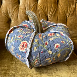 May include: A decorative, quilted pumpkin in shades of blue, red, yellow, and green. The fabric features a floral pattern and is trimmed with a white, ruffled edge. The pumpkin has a natural, brown stem.