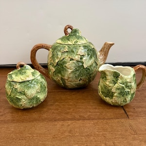 May include: A three-piece tea set featuring a teapot, sugar bowl, and creamer. The set is decorated with a green leaf pattern over a cream-colored base. The teapot has a brown handle and spout, and the sugar bowl has a matching lid.