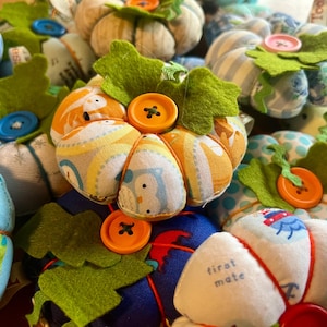 May include: A collection of fabric pumpkins with different patterns and colours. Each pumpkin has a button in the centre and green felt leaves. The pumpkins are perfect for autumn decor.
