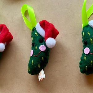 May include: Three green felt pickle ornaments with red and white Santa hats and smiling faces. Each ornament has a green ribbon for hanging and a small tag. Festive Christmas decorations.