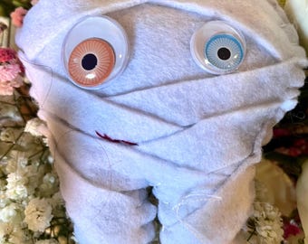 May include: A handmade, plush tooth wrapped in white fabric, with large googly eyes and a stitched red mouth. A small tag reads "tooth!" The tooth is surrounded by pink and white flowers.