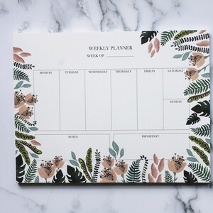 May include: A weekly planner notepad with a floral and foliage design. The notepad has a white background with black text that reads "Weekly Planner" and "Week of". The notepad is surrounded by a border of green leaves, pink flowers, and brown stems.