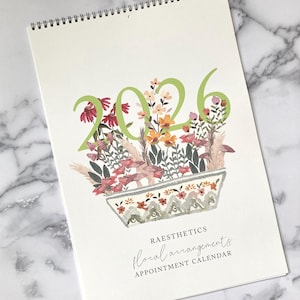 Op de afbeelding: Een spiraalgebonden afsprakenkalender voor 2026 met een bloemenontwerp. De omslag toont een aquarelillustratie van bloemen en bladeren in een decoratieve pot, met het jaar 2026 in groen. De tekst "Raesthetics Floral Arrangements Appointment Calendar" staat eronder.