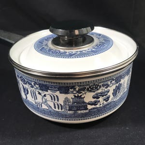 Puede incluir: Una olla de cerámica blanca y azul con tapa. La olla presenta un diseño de patrón Willow azul y blanco alrededor del exterior y en la tapa. La tapa tiene un pomo negro. La olla tiene un borde plateado y un asa negra.