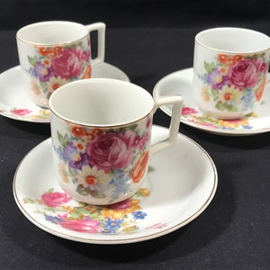 Może przedstawiać: Zestaw trzech białych porcelanowych filiżanek do herbaty i spodków z kwiatowym wzorem z różowymi, żółtymi i niebieskimi kwiatami. Filiżanki mają złoty obrzeże.