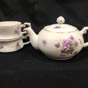 May include: A white porcelain tea set featuring a teapot and two stacked creamers. The set is decorated with purple floral patterns and gold trim. The teapot has a rounded body, a spout, and a lid with a finial.