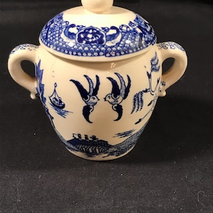 May include: A small, white ceramic sugar bowl with a lid, decorated with a blue willow pattern. The bowl has two handles and a small knob on the lid. The design includes birds, a boat, and a landscape scene.