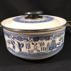 May include: A white and blue Willow pattern enamelware pot with a lid and two handles. The pot features a detailed blue landscape design around the exterior and on the lid. The lid has a black knob.