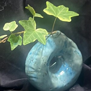 May include: A donut-shaped ceramic vase with a glossy, mottled blue and white glaze. The vase features a central circular opening and a small top aperture holding a sprig of green and yellow variegated ivy leaves with pointed lobes.
