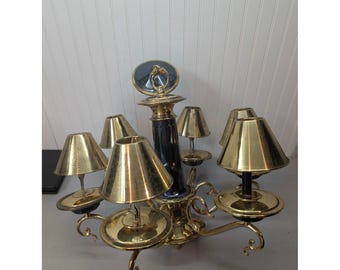May include: A brass chandelier featuring five arms with matching conical brass shades and a central dark blue column. The fixture has curved metal supports and a circular top ring, showcasing a polished metallic finish throughout the design.