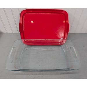 May include: A rectangular clear glass baking dish with integrated side handles and a matching solid red plastic lid. The dish features a smooth interior surface and a flat rim, while the lid is designed to fit securely over the top edges.