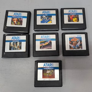 May include: A collection of seven Atari 5200 video game cartridges. The cartridges are black with white labels featuring game titles like "Missile Command," "Pac-Man," and "Space Invaders." The labels have colorful graphics and the Atari logo.