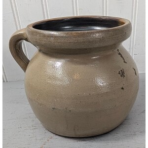 May include: A round, tan-colored stoneware crock with a single side handle and a wide, thick rim. The exterior has a textured, glazed finish with visible dark brown spots and imperfections. The interior is dark brown and smooth in appearance.