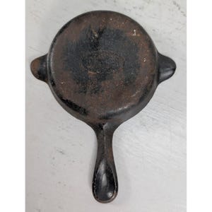 May include: A small, round, cast iron skillet with a long handle and two small side handles. The skillet is dark brown with signs of wear and tear. The text "WAGNER" and "MADE IN USA" are visible on the bottom of the skillet.