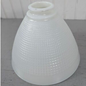 May include: A white milk glass lamp shade with a bell-shaped silhouette and a textured grid pattern across the exterior surface. The shade features a smooth, circular opening at the top for mounting and a wide, flared base for light diffusion.