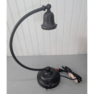 May include: A black desk lamp with a bell-shaped shade and a curved arm. The lamp has a round base and a power cord. The lamp is made of metal and has a matte finish. The lamp is designed for desk use.