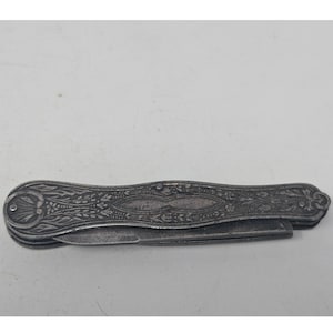 May include: A closed pocket knife with a metallic silver-toned handle featuring an intricate embossed floral and scrollwork pattern. The handle has a curved, elongated shape with a rounded end and a visible metal pin near the pivot point.