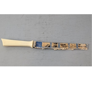 May include: A kitchen knife with a cream-colored plastic handle and a blade encased in a blue cardboard sheath. The sheath features five small black and white illustrations demonstrating the use of the knife and has frayed, worn edges.