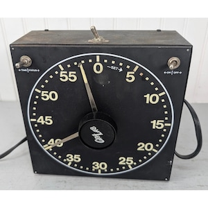 May include: A black square GraLab darkroom timer with a large circular face featuring white numbers from 0 to 55. It includes a central black control knob, two metal toggle switches labeled Time/Focus and On/Off, and a top toggle switch.