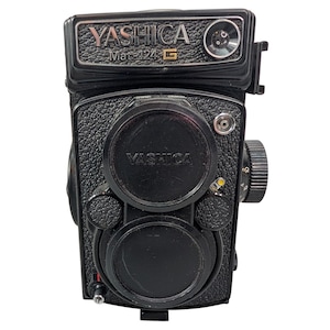 May include: Black Yashica Mat-124 G camera with the brand name on the top and lens cover. The camera has a textured body, a lens cap, and a dial on the side. The camera is a vintage model.