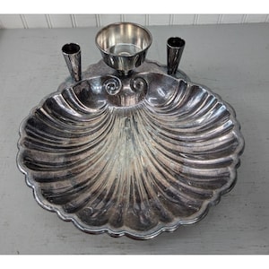 May include: A silver-toned metal desk accessory shaped like a large scallop shell. It features a central circular bowl and two smaller cylindrical holders on either side, all integrated into the shell-shaped base with ribbed detailing.