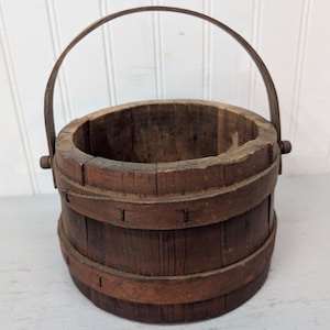 May include: A small, antique wooden bucket with a dark brown finish. The bucket has a metal handle and is held together with wooden bands. The wood grain is visible, giving it a rustic appearance. The bucket is empty.