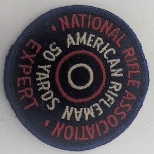 May include: A circular embroidered patch with a dark blue background and a red and beige target design in the center. The text reads NATIONAL RIFLE ASSOCIATION, AMERICAN RIFLEMAN, 50 YARDS, and EXPERT in a mix of red and beige stitched lettering.