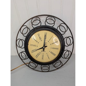 May include: A vintage wall clock with a black metal frame and a cream-colored face. The clock features black Roman numerals and hands, with a decorative circular frame. The clock is powered by a cord.