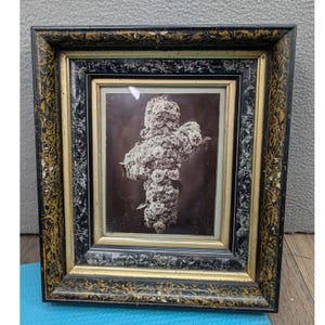May include: A framed sepia-toned photograph of a cross made of flowers. The frame is black with gold accents. The cross is centered in the frame and is made of white and light-colored flowers. The background is a dark brown color.