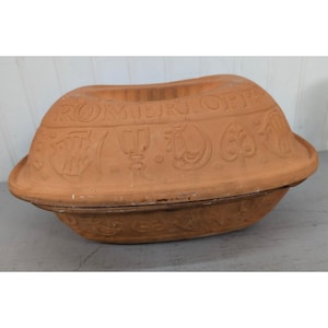 May include: A terracotta Romertopf clay baker with a lid. The exterior features embossed designs including a fish, a chalice, a rooster, and a leaf motif. The word ROMERTOPF is clearly stamped across the top section of the lid in capital letters.