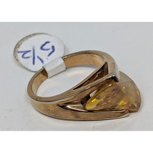 May include: A gold-toned ring with a large, triangular, yellow gemstone. The ring has a split shank design and a setting that cradles the gemstone. A small tag with handwritten text is attached to the ring.