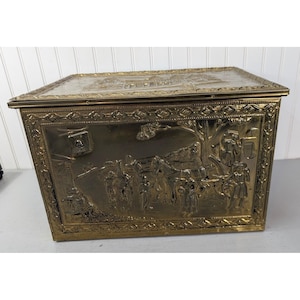 May include: A rectangular brass storage box featuring an embossed scene of figures, a horse-drawn carriage, and trees on the front panel. The lid and edges are decorated with intricate geometric patterns, and a small metal latch is on the left side.