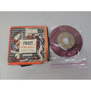 May include: An album cover and a vinyl record. The album cover is orange and features illustrations of figures in a theatrical setting. The record is a deep red color and is in a clear plastic sleeve. The album is titled "Faust".