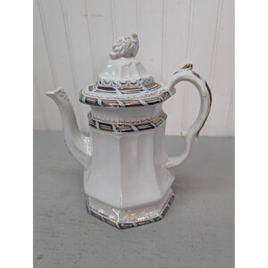 May include: A white porcelain teapot with an octagonal body and a matching lid featuring a sculpted finial. The teapot is decorated with horizontal bands of metallic gold, blue, and purple trim along the lid, the upper rim, and the base.