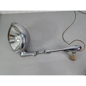 May include: A vintage chrome spotlight with a reflective interior and adjustable arm. The spotlight has a bulb and a wire extending from the base. The light is mounted on a gray surface.