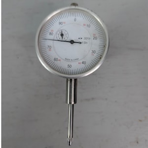 May include: A silver dial indicator with a white face and black markings. The dial reads from 0 to 100, with smaller increments. The text "MADE IN CHINA" is visible. The indicator has a long, silver stem.