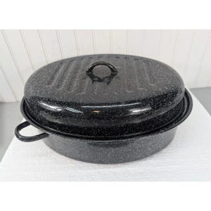 May include: An oval-shaped black roasting pan with a white speckled enamel finish. The pan features a matching lid with a central handle and two side handles on the base. The lid has a series of vertical ridges across its surface.