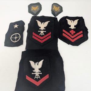 May include: A collection of black fabric military patches featuring embroidered designs. The set includes two small eagle patches, one patch with a star and crosshair, and three larger shield-shaped patches displaying eagles, red chevrons, and symbols.