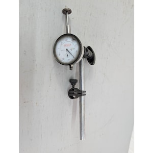 May include: A dial indicator gauge with a white face, black numbers, and a black needle. The gauge is mounted on a metal stand with a black clamp and a silver rod. The gauge measures in inches.