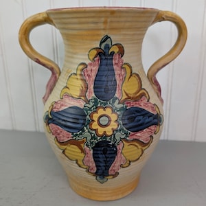 May include: A cream-colored ceramic vase with two handles and horizontal ribbed texture. The exterior features a central hand-painted floral motif in shades of blue, yellow, pink, and green, with a symmetrical design radiating from a central flower.