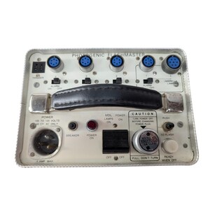 May include: A vintage Photogenic Flashmaster unit with a silver metal casing, black handle, and various buttons and ports. The unit has text labels including "Power On," "Trigger," and "Caution." It features multiple blue circular ports.