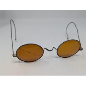 May include: Antique oval-shaped sunglasses with amber-colored lenses and a thin, silver-toned metal frame. The lenses show a crack. The temples are wire-like and curve over the ears. The glasses are set against a white background.
