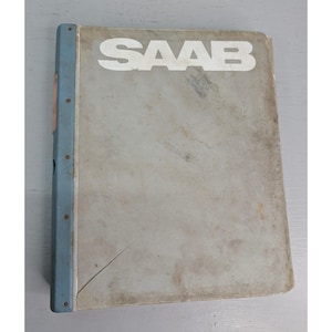 May include: A vintage SAAB manual with a light blue spine and the word "SAAB" in white block letters on the cover. The cover is a faded gray color with some wear and tear.