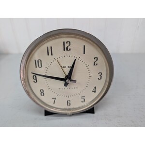 May include: A round Westclox Big Ben alarm clock with a silver-toned metal frame and a cream-colored face. The clock features black Arabic numerals, black hour and minute hands, a thin gold-toned second hand, and a textured dial surface.