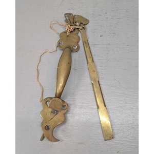May include: A brass door handle set featuring a curved grip with ornate, scalloped mounting plates at each end, accompanied by a long, flat, rectangular brass arm. The two components are connected by a thin, light-colored string.