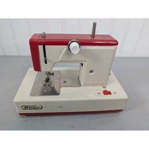 May include: A vintage off-white and red sewing machine with the brand name "Strace" printed on the front. The machine has a red top and a silver needle plate. The on/off switch is red. The machine is on a gray surface.