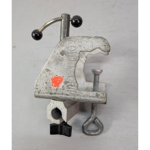 May include: A silver-colored metal clamp with a screw and handle. The clamp has a black handle and a red sticker. The clamp is designed to hold objects in place. The word "GRIP" is visible on the side of the clamp.
